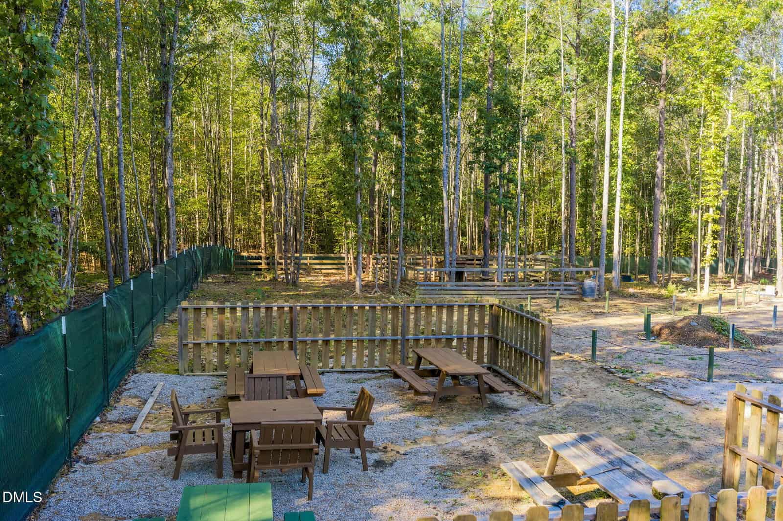 Outdoor area with wooden picnic tables and benches enclosed by a low wooden fence, surrounded by trees and a green mesh fence, with gravel and dirt ground visible. The setting appears to be in a forested area.