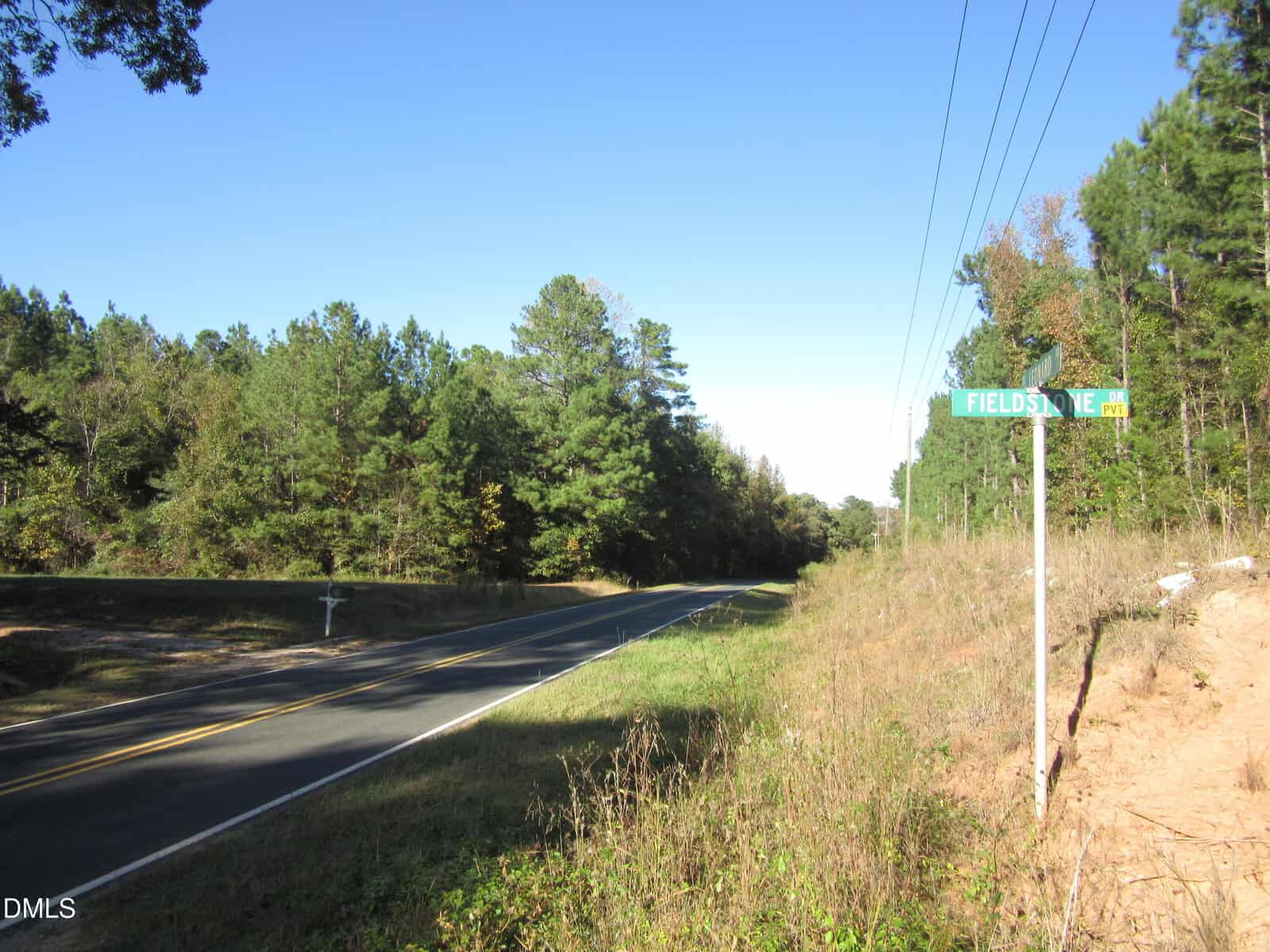 A rural two-lane road with a yellow centerline curves beside trees and grass near Louisburg. A green street sign reads “FIELDSTONE CT.” Power lines run along the right side under a clear blue sky, bordering the 4.29 acres on Leonard Road.