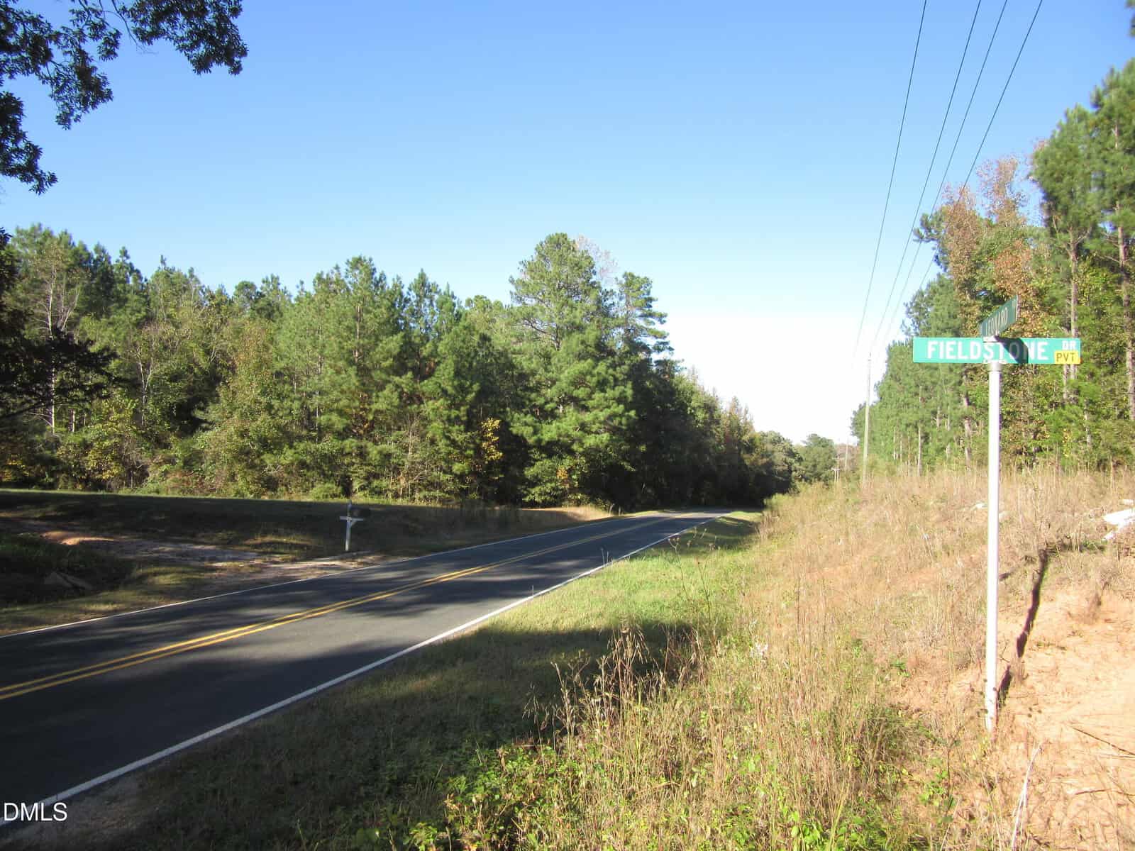 A two-lane road curves through a rural area with trees on both sides. A green street sign reads “Fieldstone Dr.” Power lines run alongside the road under a clear blue sky.