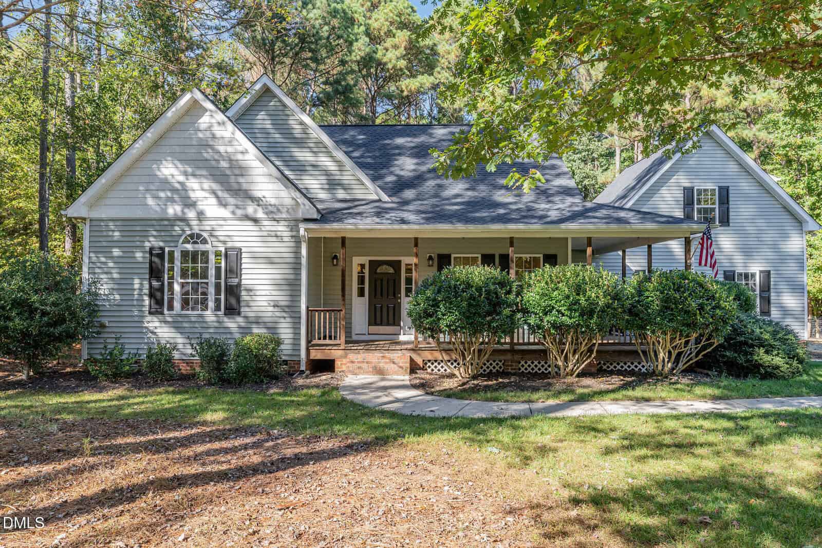 A light gray house with white trim and a covered front porch sits among trees. There are shrubs along the porch, a brick entryway, and an American flag hanging near a side entrance.