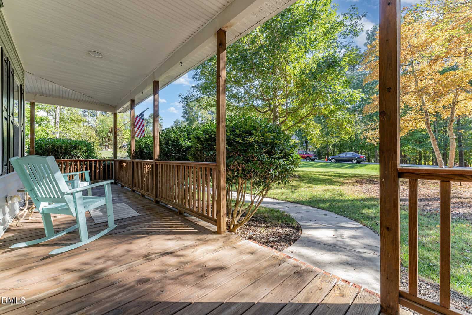 A wooden front porch with two light blue chairs faces a curved concrete walkway. The porch has a wooden railing, nearby bushes, and trees in the background. An American flag hangs near the house.
