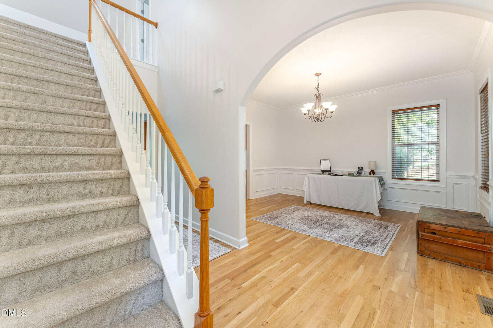 A carpeted staircase with a wooden handrail is on the left, leading up to a second floor. On the right, there is a room with hardwood floors, a rug, a table with a laptop, a chandelier, and windows with blinds.