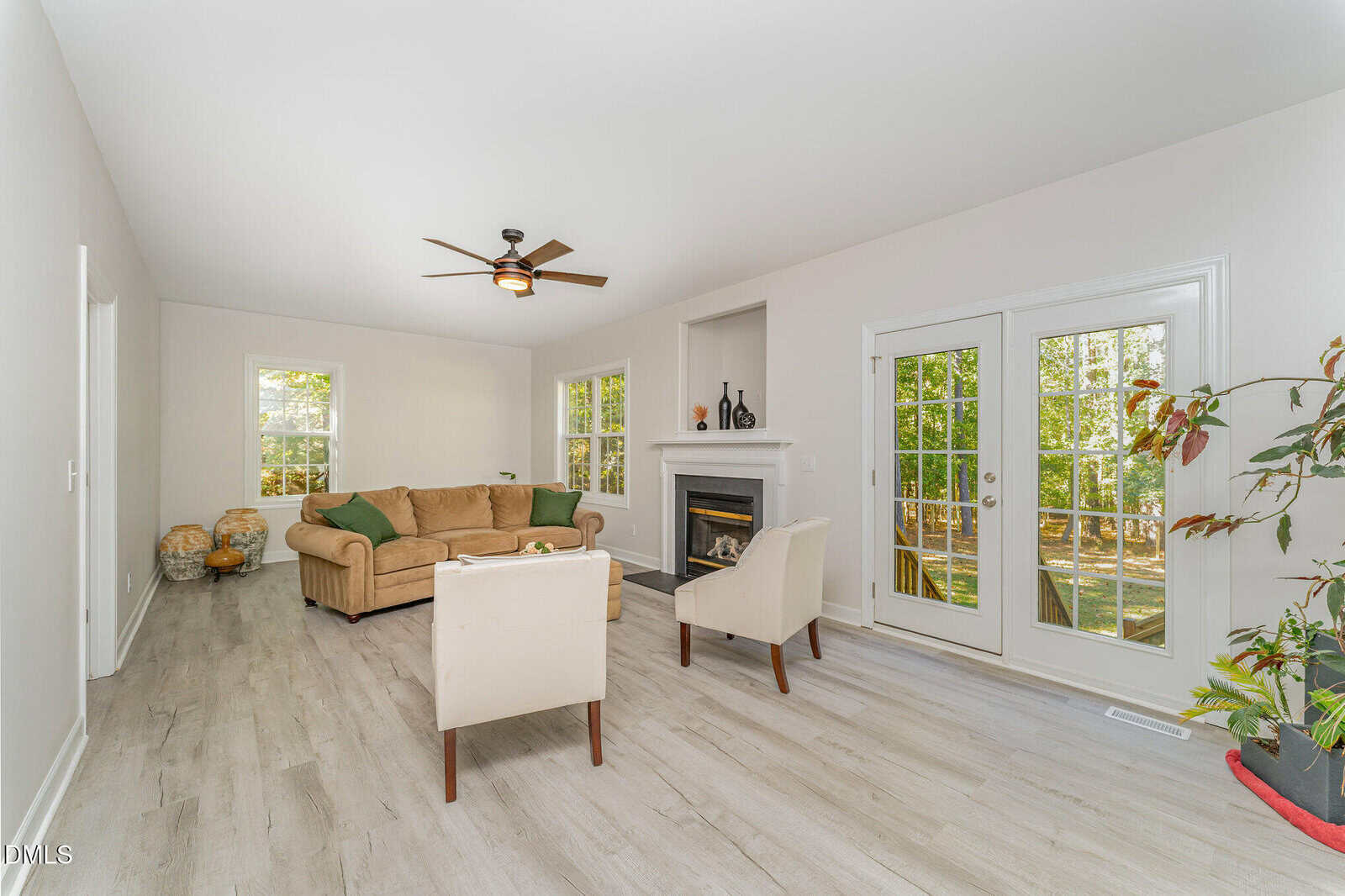 A bright living room with light wood flooring, beige sofa, two white armchairs, a fireplace, ceiling fan, and double glass doors opening to a yard. The room has white walls and large windows.