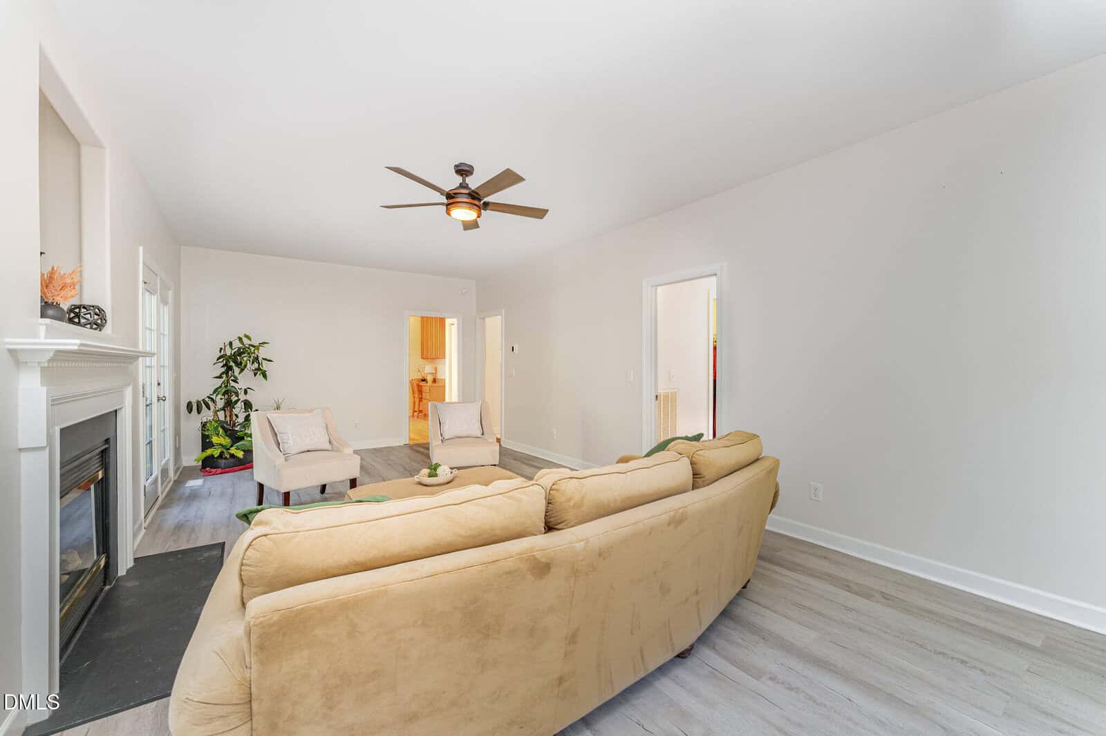A living room with light-colored walls, a beige sectional sofa, two cream chairs, a fireplace on the left, a ceiling fan, a potted plant, and light wood flooring. Doorways lead to other rooms in the background.