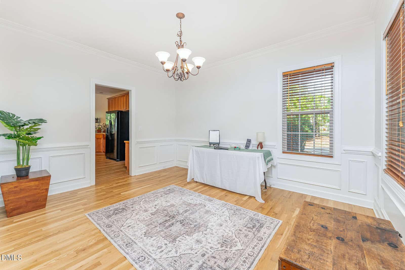 A bright, minimally decorated room with hardwood floors, an area rug, a table with a computer setup, a wooden chest, a potted plant, two windows with blinds, and a chandelier. The kitchen is visible through an open doorway.