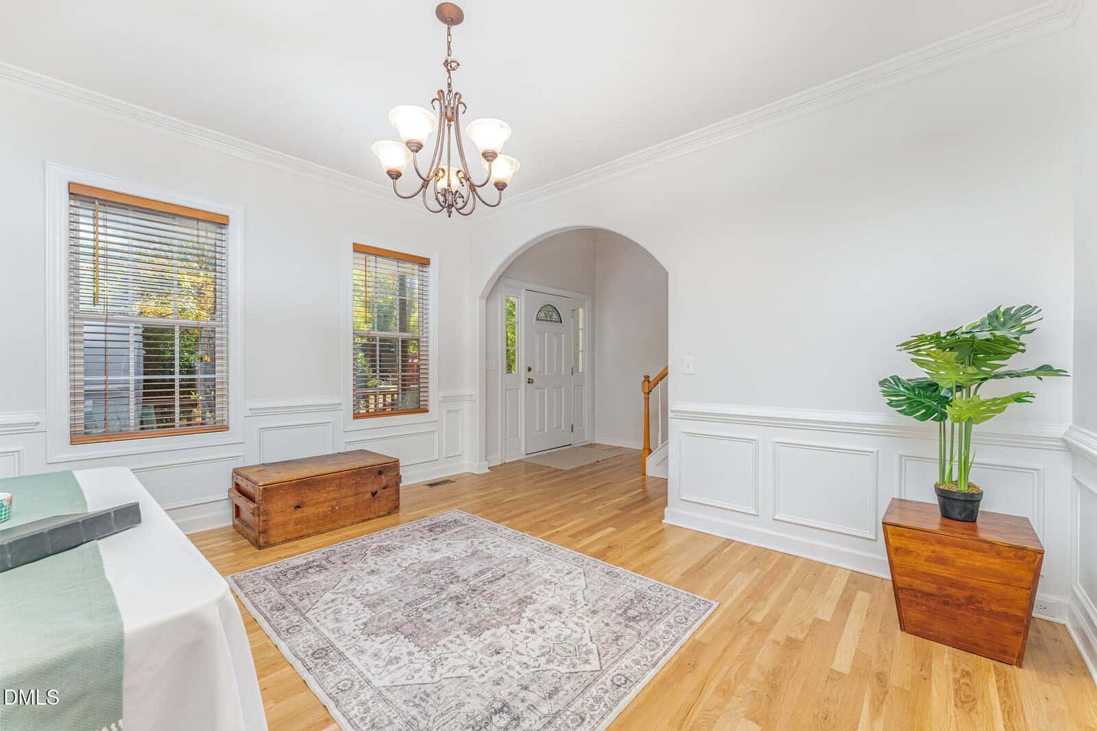 A bright entryway with hardwood floors, white walls, a patterned area rug, a wooden chest, a chandelier, a potted plant, and windows with wooden blinds letting in natural light.