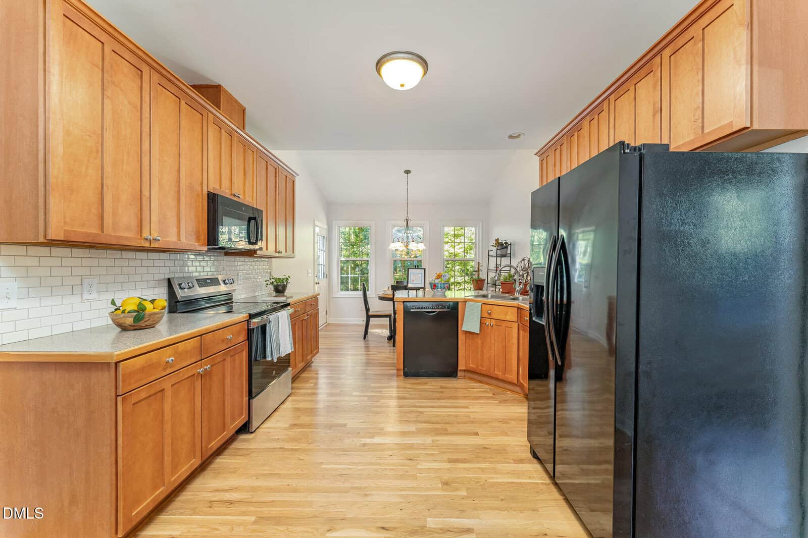 A kitchen with light wooden cabinets, light countertops, a black refrigerator, black appliances, hardwood floors, and white subway tile backsplash. A dining area with windows is visible in the background.