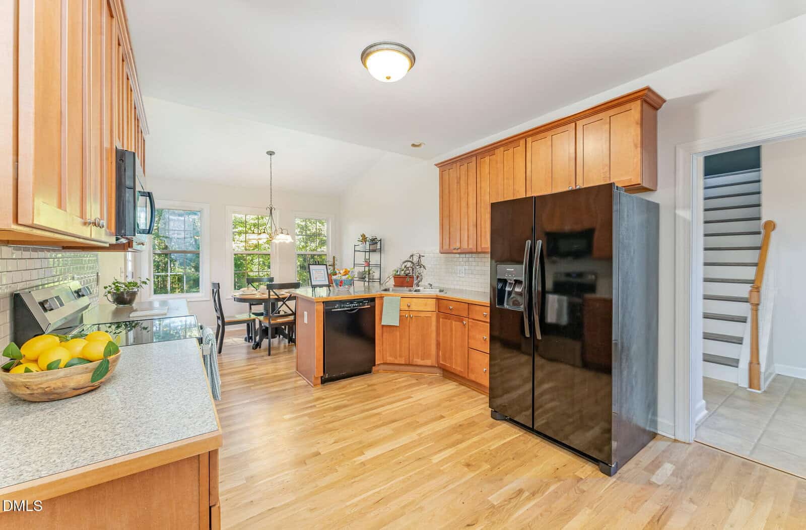 A spacious kitchen with wood cabinets, light granite countertops, black refrigerator, dishwasher, stove, and a small dining area with windows. Light wood flooring and a staircase visible in the background.