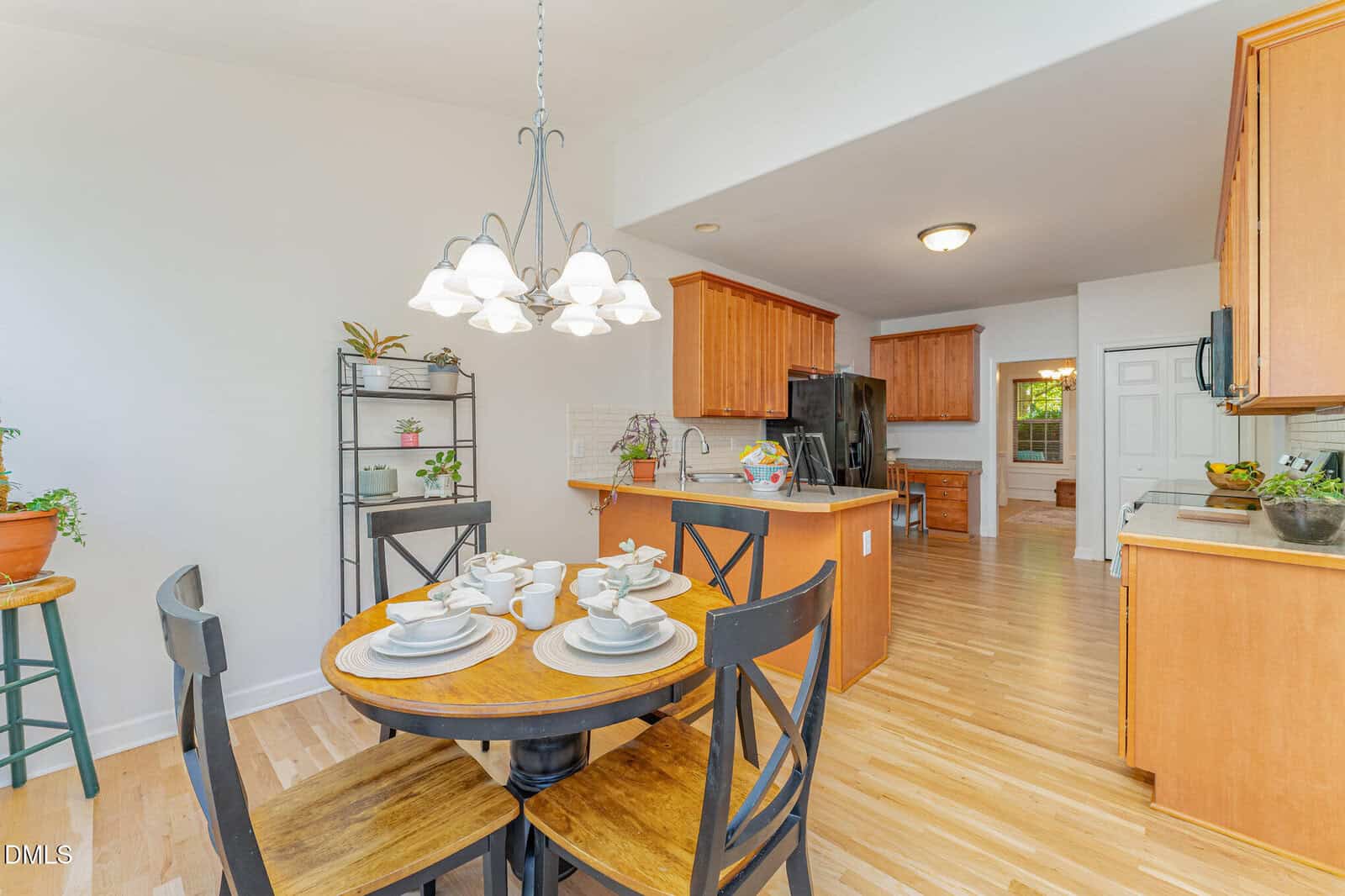 Bright kitchen and dining area with a round table set for four, wooden cabinets, black appliances, potted plants, and light wood floors. A hanging light fixture is above the dining table.
