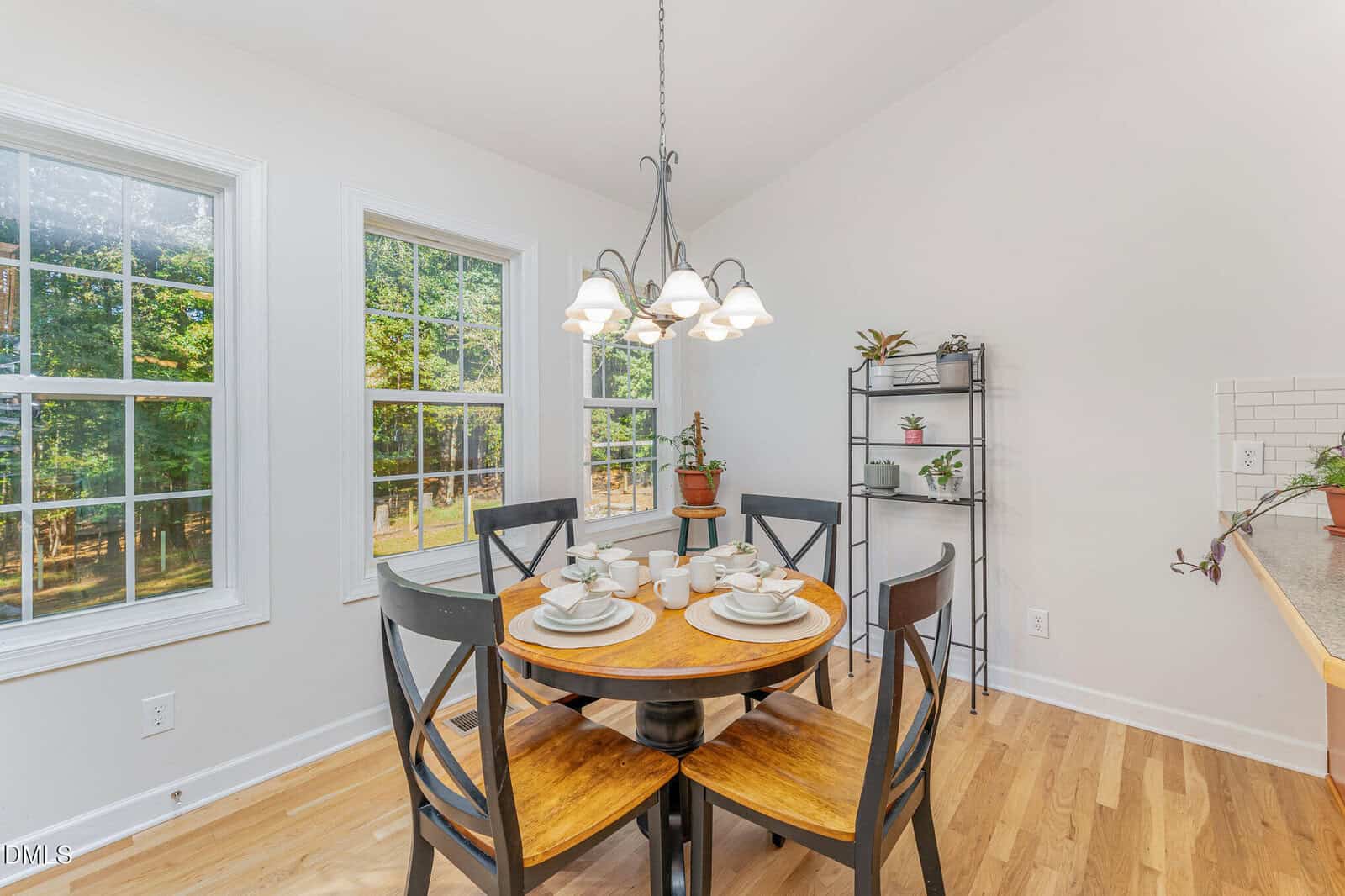 A bright dining area with a round wooden table set for four, black chairs, large windows letting in sunlight, a hanging light fixture, and a black metal plant shelf by the wall.