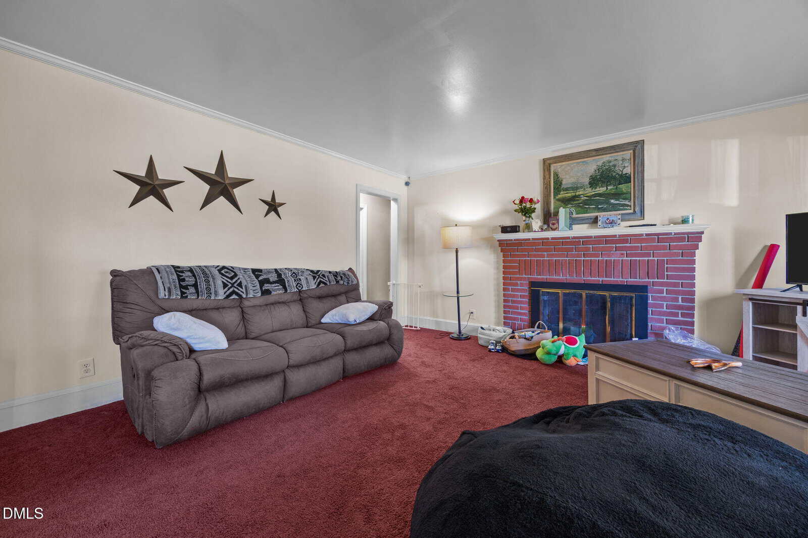 A living room with a brown sofa against a cream wall decorated with three metal stars, a brick fireplace with a painting and small items on the mantel, a bean bag, toys on the floor, and red carpet.