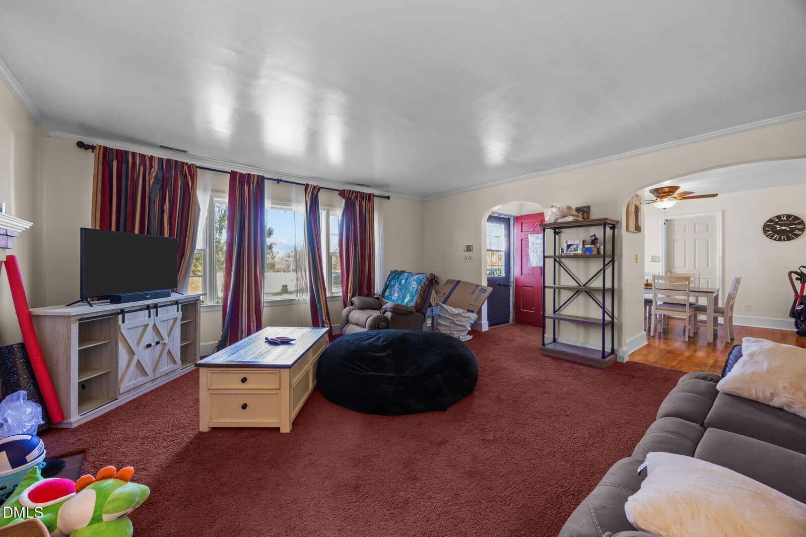A living room with red carpet, beige walls, a TV on a stand, a coffee table, a large black bean bag, a couch, and armchairs. There are large windows with purple curtains and an open view to a dining area.