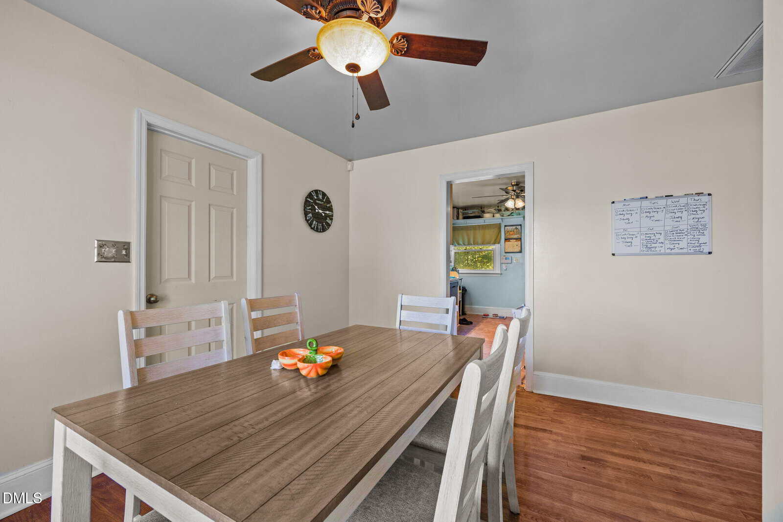 Dining room with a wooden table, six white chairs, ceiling fan, wall clock, whiteboard with notes, and fruit centerpiece. Doorway leads to another room with a green accent wall and wall art.