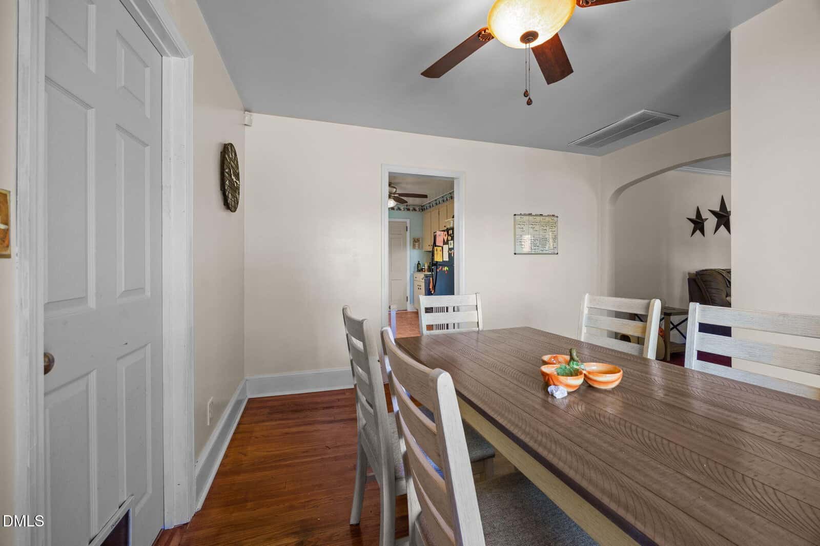 A dining room with a wooden table and six white chairs, ceiling fan above, hardwood floors, and a view into a kitchen and living room with wall decor. Three bowls and a small plant sit on the table.