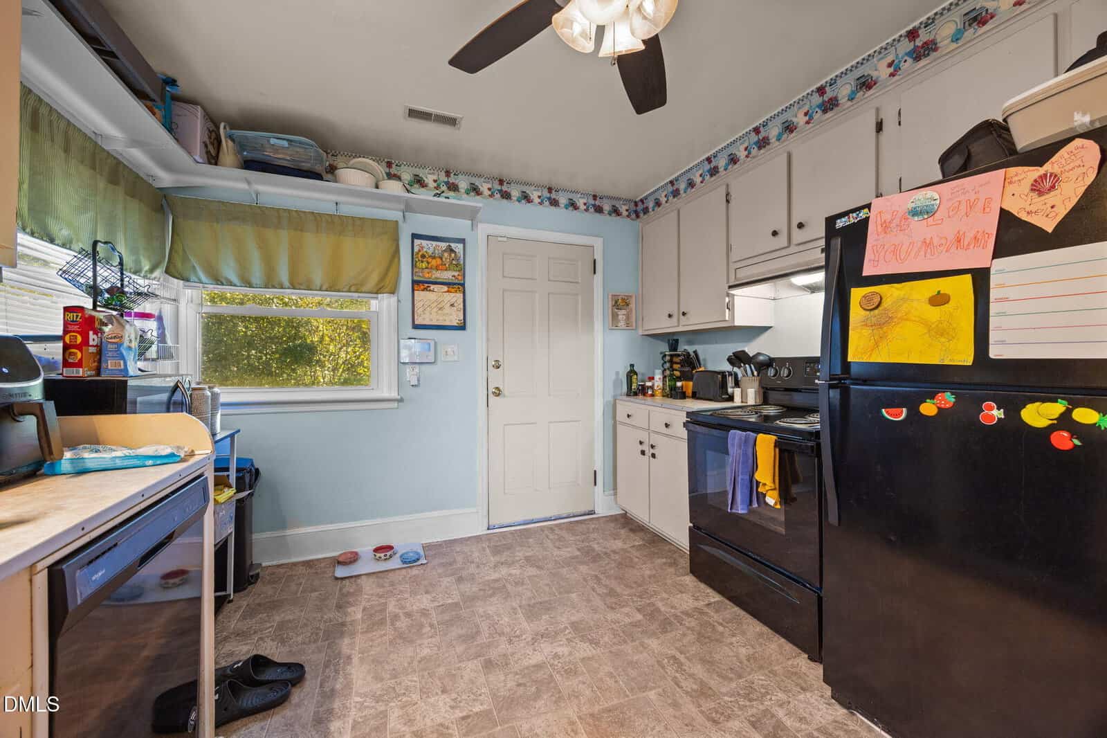 A cozy kitchen with white cabinets, black appliances, colorful kids’ drawings on the fridge, a ceiling fan, a window with green curtains, and pet bowls on the floor near the back door.