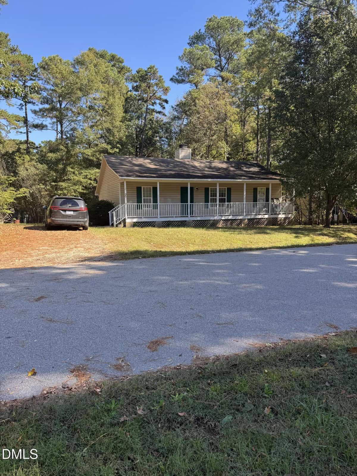 A small, single-story yellow house with a front porch and green shutters sits among tall trees. A gray car is parked in the driveway, and the house faces a quiet, paved road under a clear blue sky.