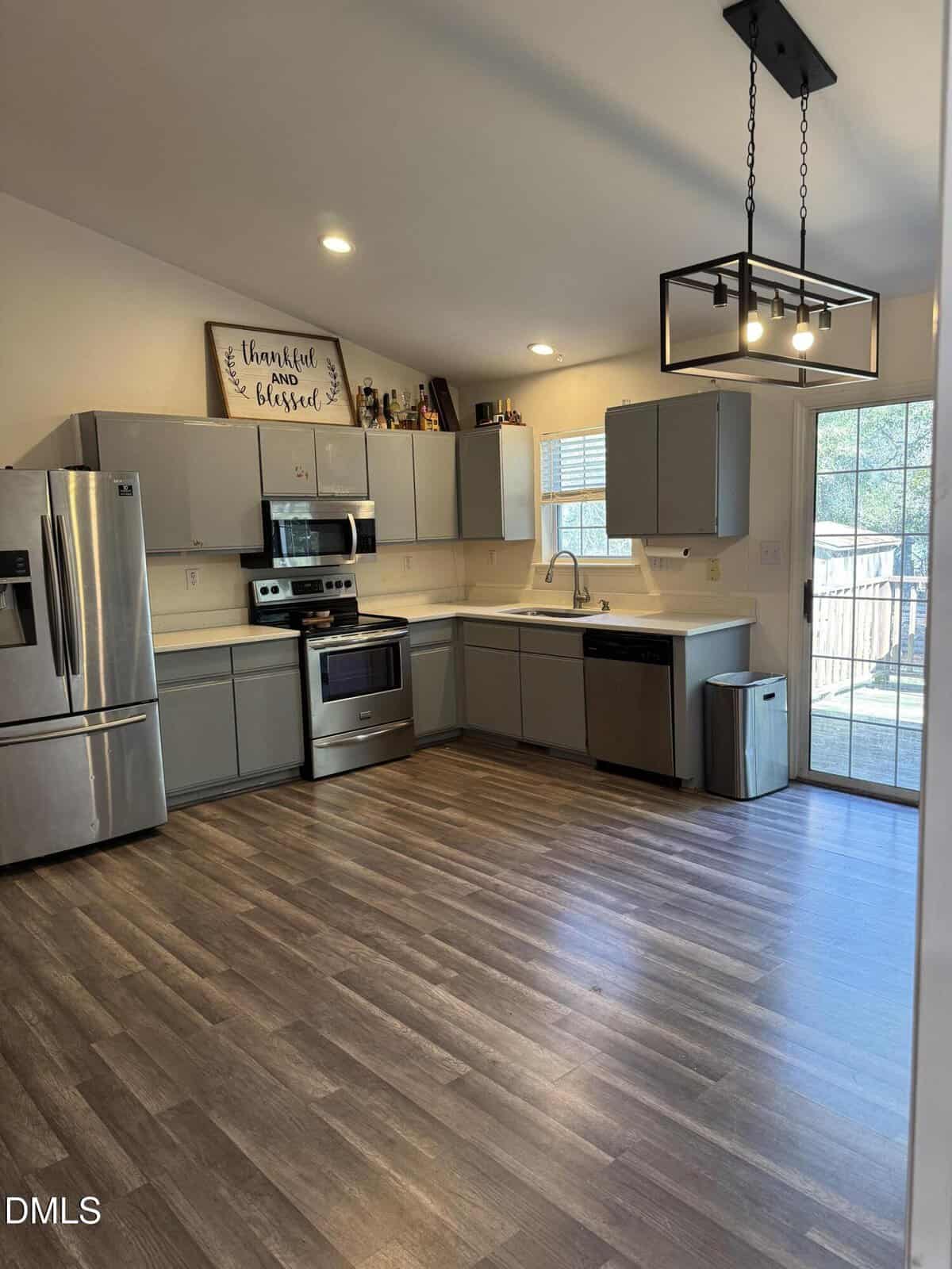 Modern kitchen with gray cabinets, stainless steel appliances, wood-look flooring, a large window and glass doors leading outside, and a decorative sign above the cabinets. Light fixture hangs from the ceiling.