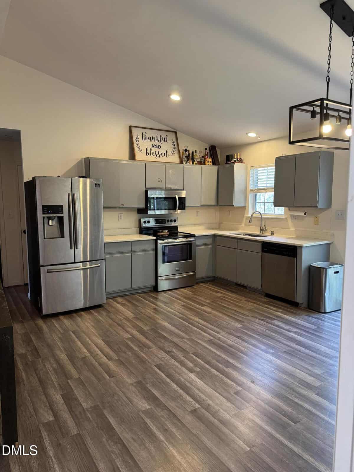 Modern kitchen with gray cabinets, stainless steel appliances, wood-style flooring, a large fridge, a stove, and a wall sign reading “thankful and blessed” above the cabinets. Natural light comes through a window over the sink.