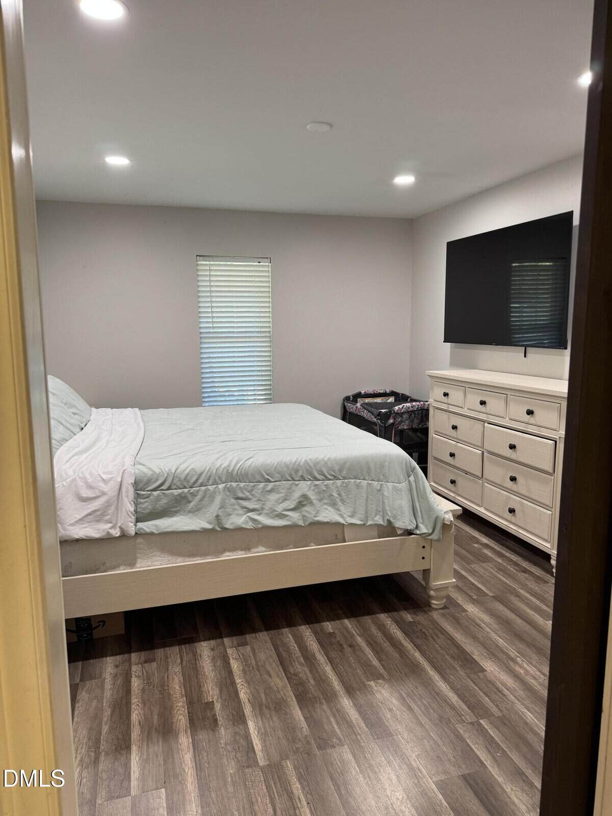 A simple bedroom with a bed, white dresser, large wall-mounted TV, window with blinds, and a playpen in the corner. The room has wood flooring and soft overhead lighting.