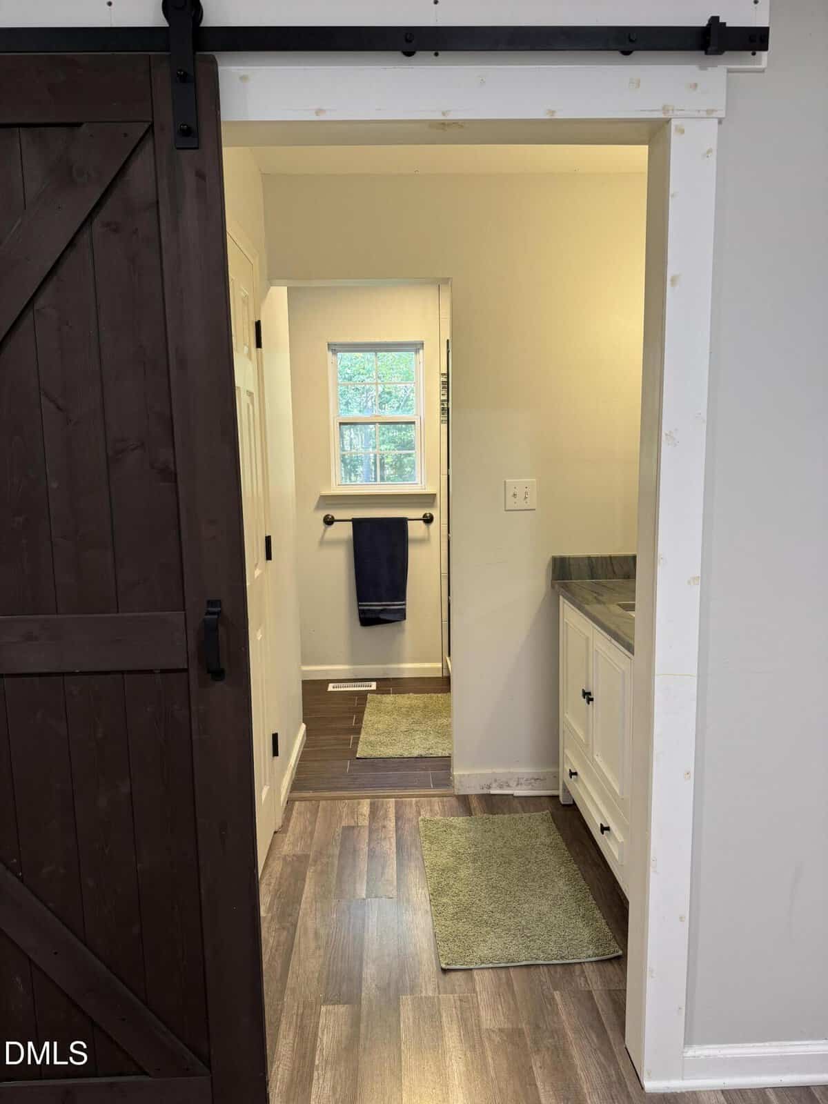 View into a bathroom with wood-style flooring, a dark sliding barn door on the left, white vanity with a dark counter on the right, two green rugs, and a window above a towel bar in the back.