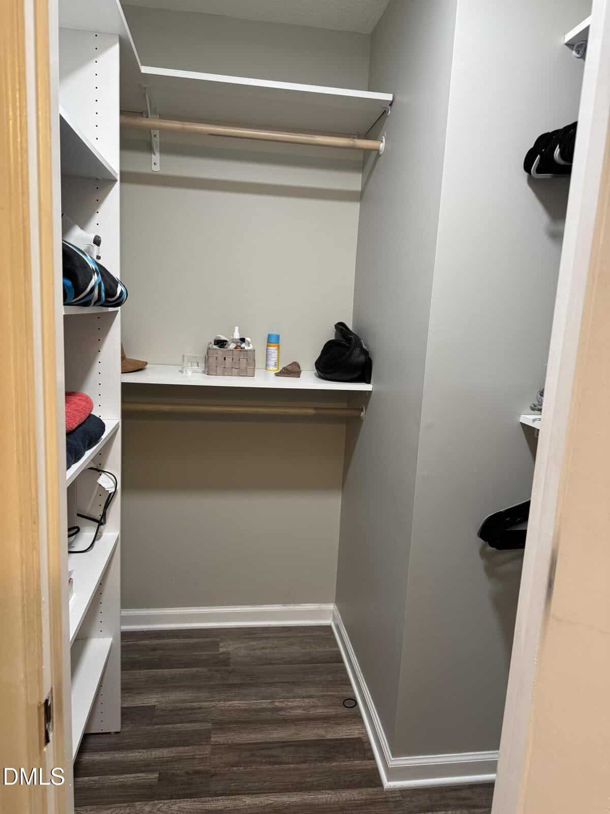A small walk-in closet with light gray walls, open shelving on the left holding folded clothes, a hanging rod, and a white shelf at the back with toiletries, baskets, and a few bags. The floor is wood-patterned laminate.