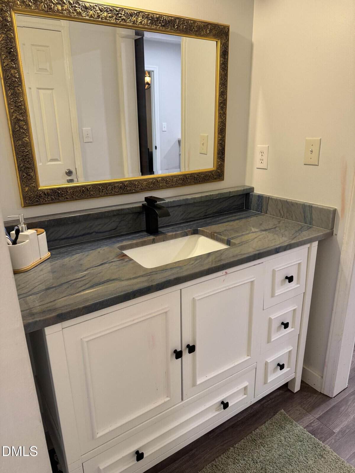 A bathroom vanity with a gray marble countertop, white cabinetry, black faucet, and a rectangular mirror with a gold ornate frame above it. Toiletries sit on the counter and a green bath mat is on the floor.
