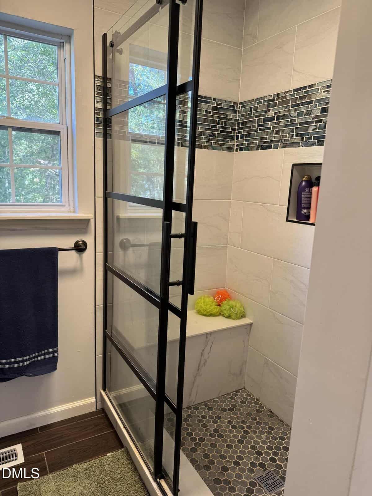 Modern shower with a glass door featuring black frames, a built-in bench, blue tile accent, and shelves with toiletries. A towel hangs nearby; a window lets in natural light.