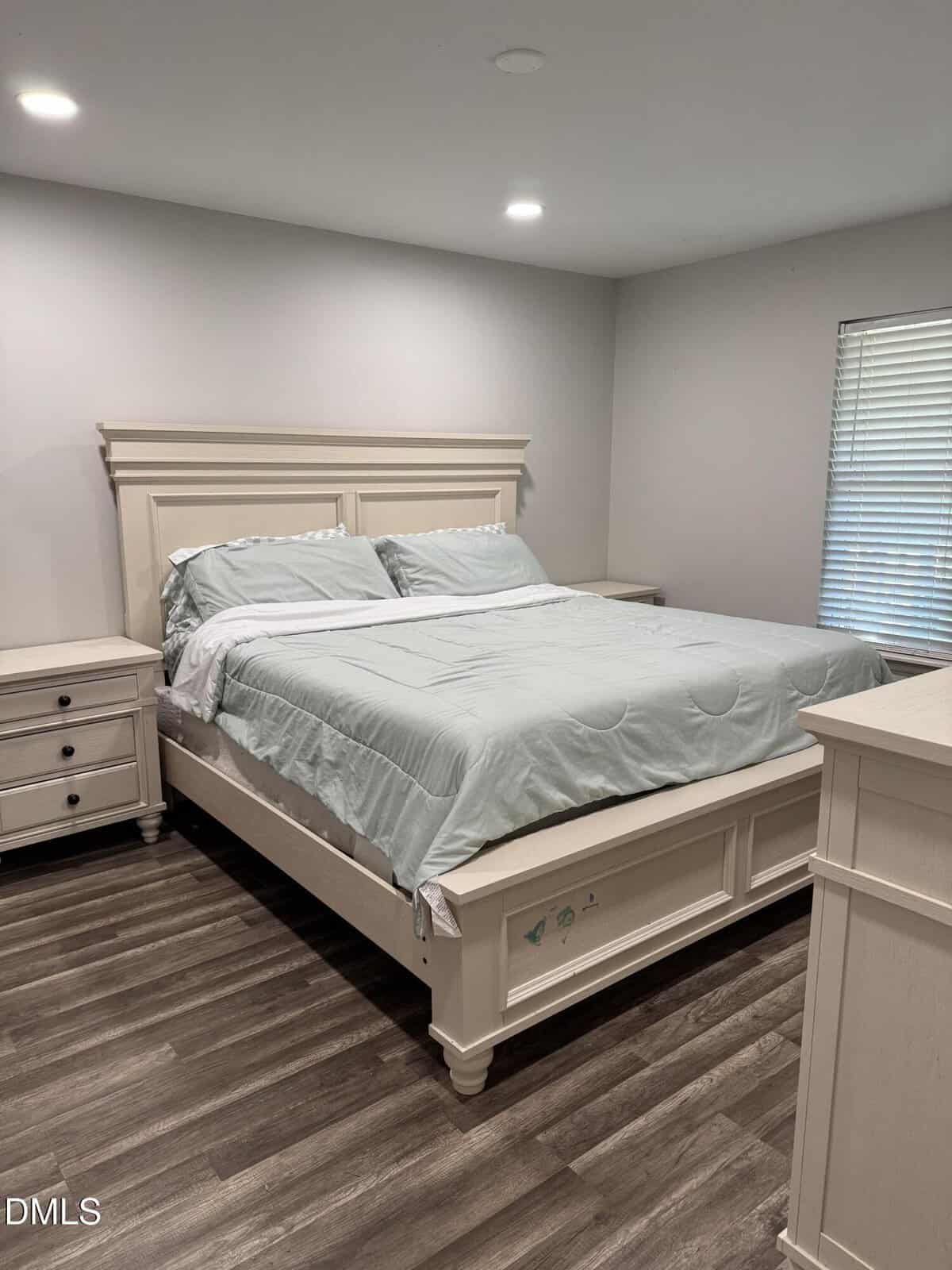 A neatly made queen-size bed with light blue bedding sits between two white nightstands in a modern bedroom with gray walls, wood floors, and a window with closed blinds on the right.