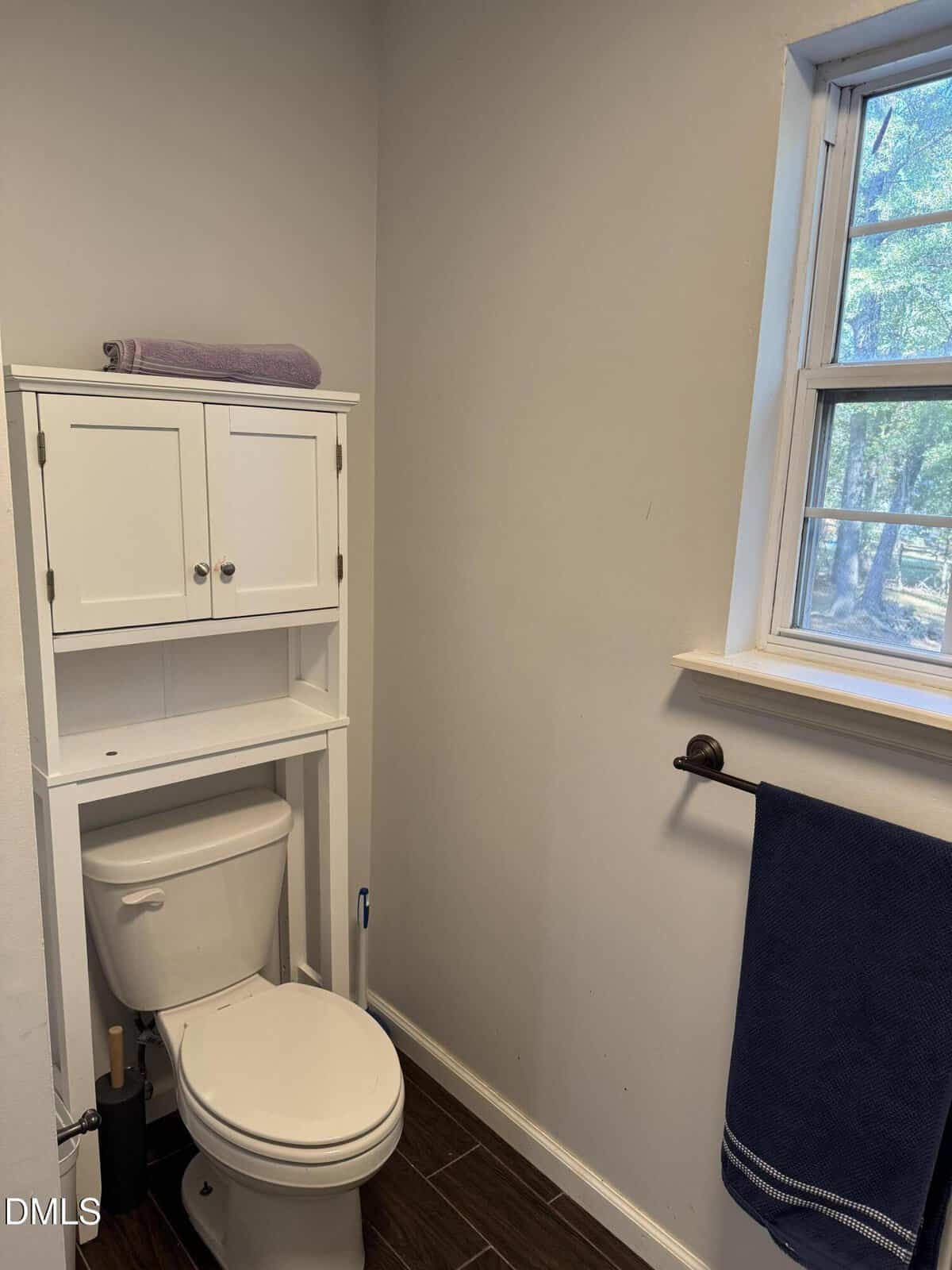 A small bathroom with a white toilet under a white cabinet, a purple towel on top, a window with a view of trees, and a dark blue towel hanging on the wall. The floor has dark wood-like tiles.
