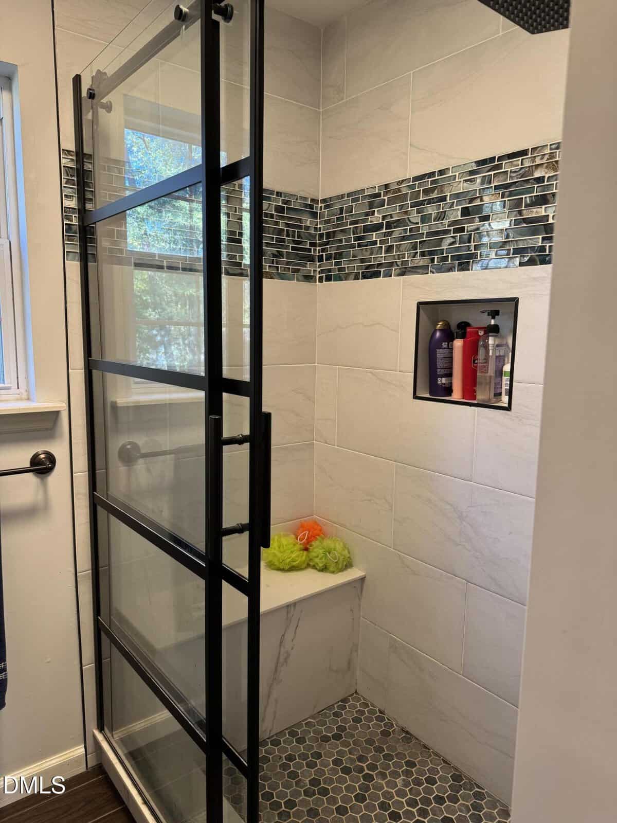 Modern shower with glass door, white tile walls, blue mosaic accent strip, built-in shelf holding bottles, and a bench with a green bath puff and orange container. Hexagonal floor tiles and window visible.