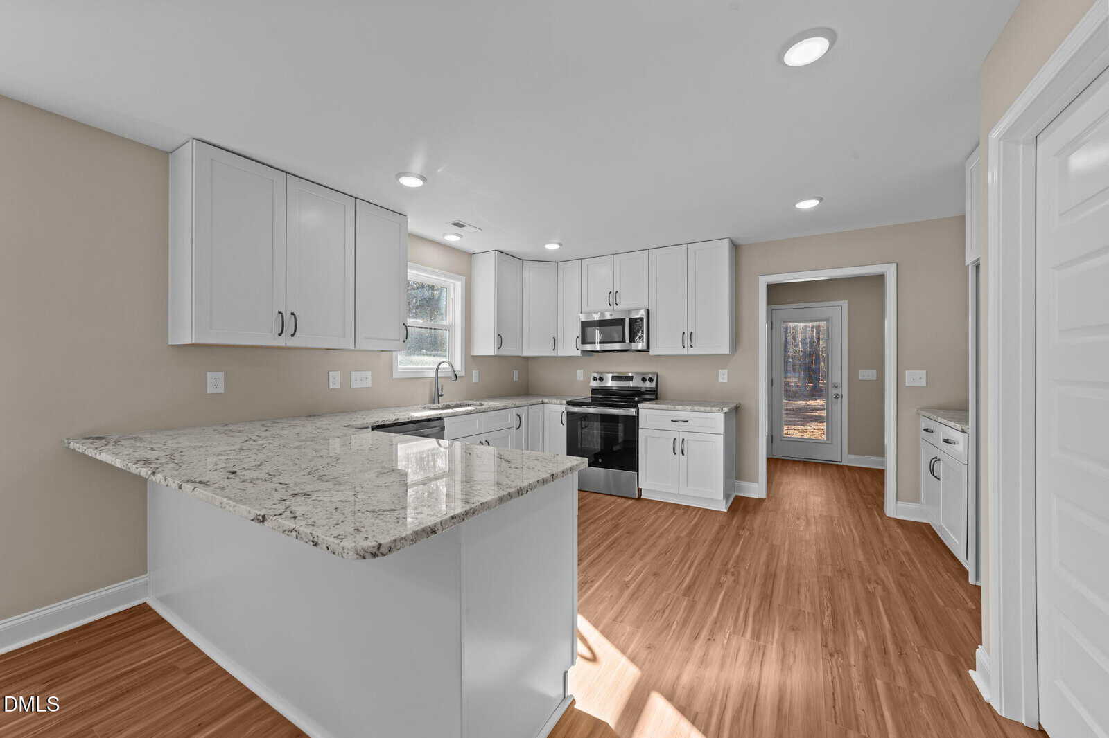 Modern kitchen with white cabinets, stainless steel appliances, light granite countertops, and a large island. The room has wood flooring, beige walls, and a window above the sink letting in natural light.