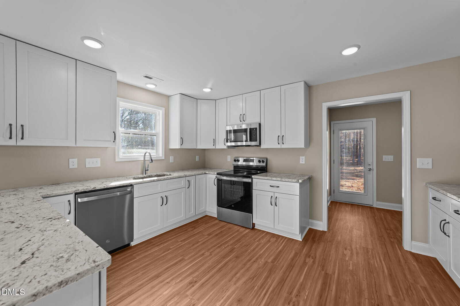 Modern kitchen with white cabinets, stainless steel appliances, marble countertops, light wood flooring, and a window above the sink. A doorway leads to another room with glass doors and a view of trees outside.