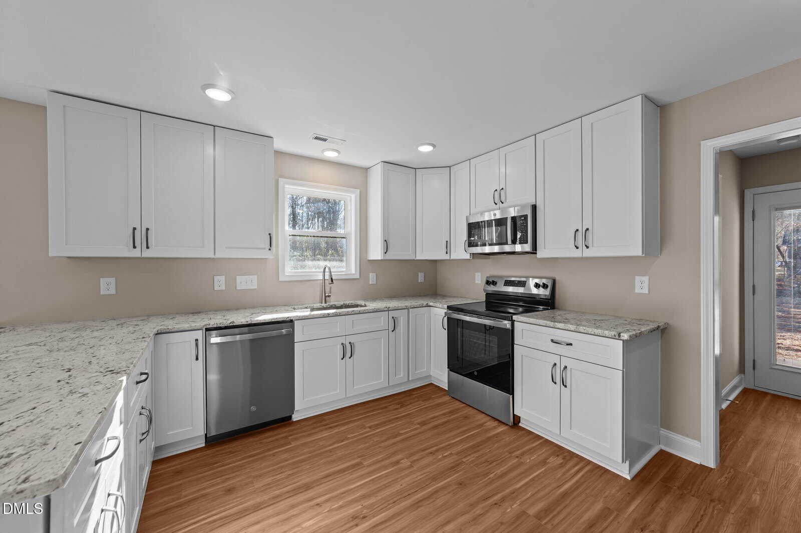 Modern kitchen with white cabinets, stainless steel appliances, marble countertops, wood flooring, and a window above the sink letting in natural light.