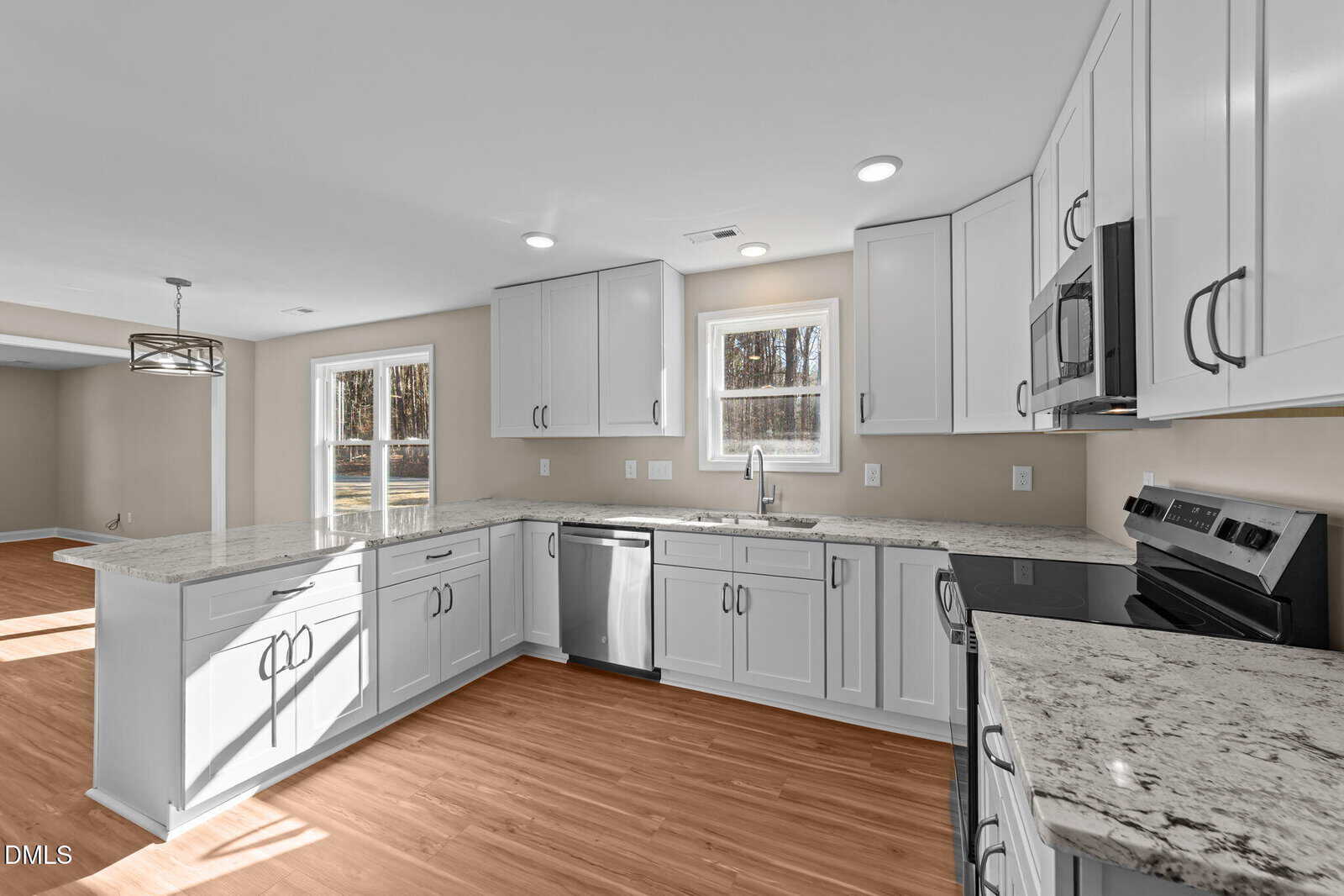 Bright modern kitchen with white cabinets, granite countertops, stainless steel appliances, and wood-style flooring. Large windows let in natural light and overlook an open dining area.