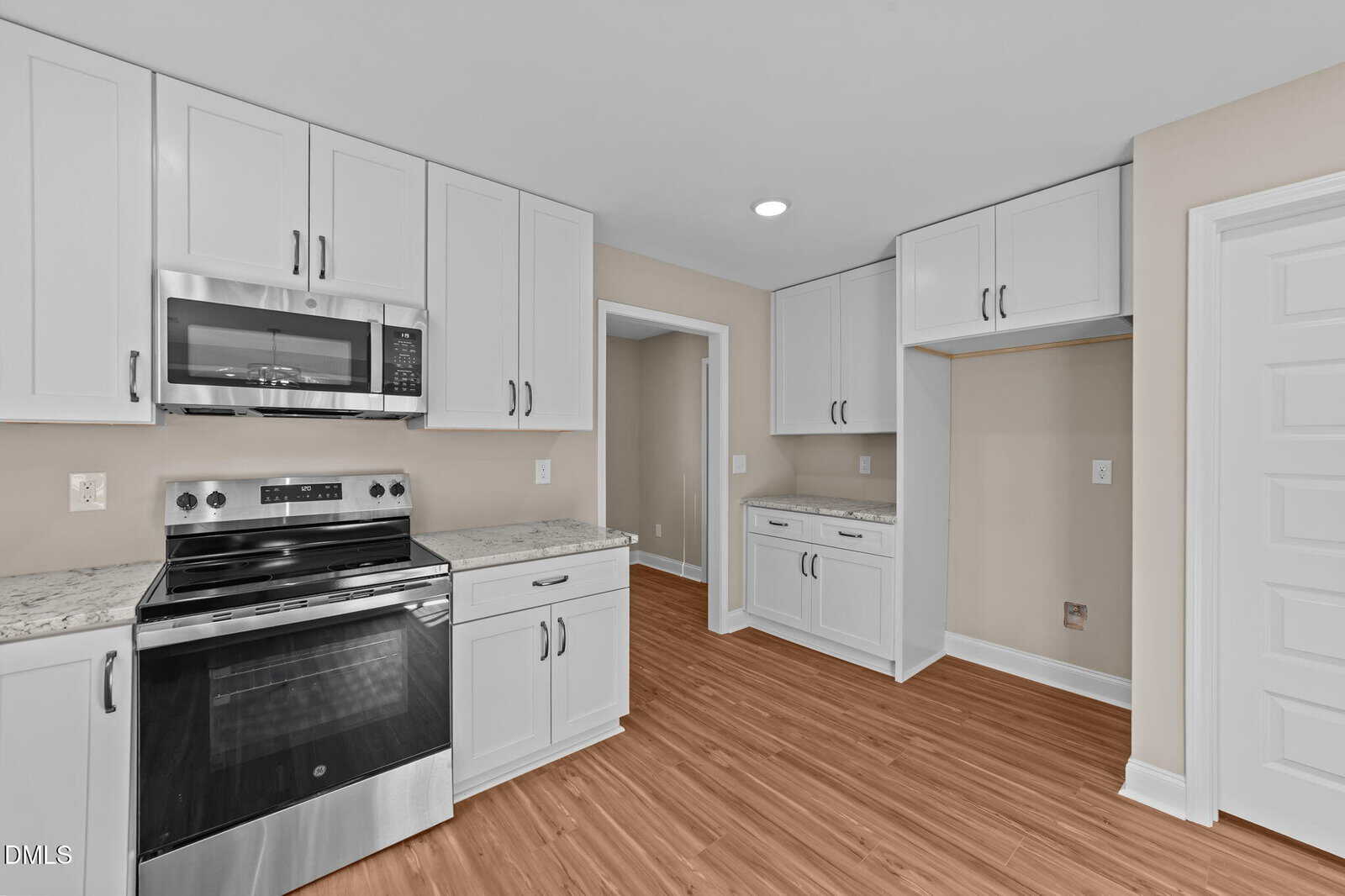 Modern kitchen with white cabinets, stainless steel oven and microwave, light granite countertops, beige walls, wood-look flooring, and built-in pantry space. Room is well-lit with recessed lighting.