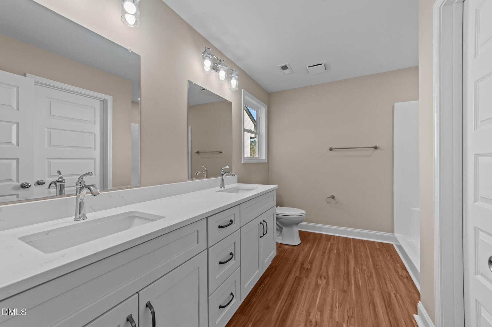 Modern bathroom with a double-sink white vanity, two mirrors, wood-look flooring, toilet, towel bar, a window, and a shower. Neutral color palette with bright lighting.