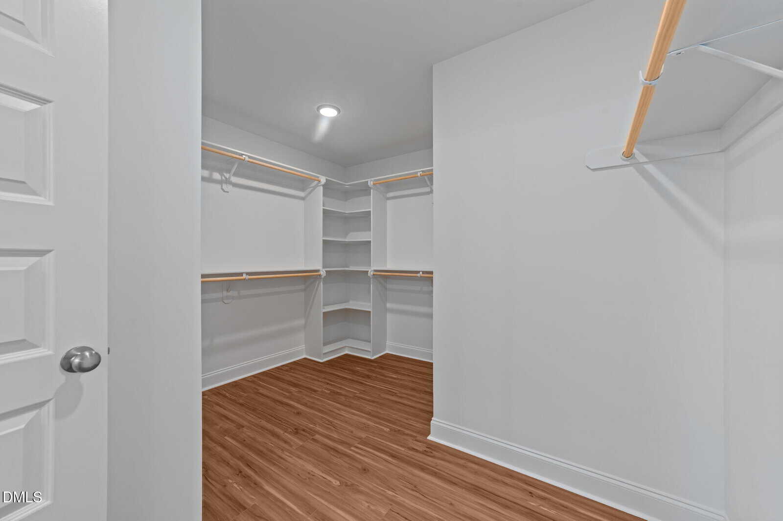 Spacious empty walk-in closet with white walls, built-in shelves, and wooden hanging rods. The floor is light brown wood, and the room is well-lit by a ceiling light.