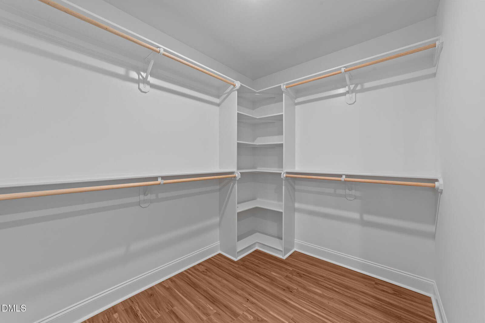 Spacious, empty walk-in closet with white walls, wooden rods for hanging clothes, built-in corner shelves, and wood-patterned flooring.