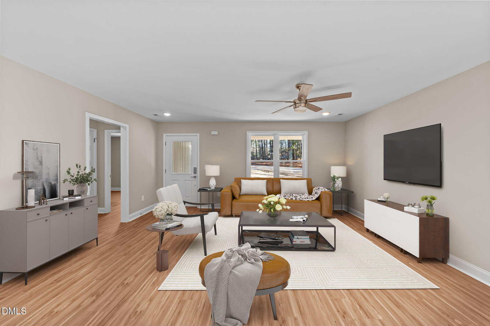 A modern living room with light wood flooring, beige walls, a brown sofa, a white rug, a round ottoman, and a wall-mounted TV. Large windows let in natural light, and minimalist furniture decorates the space.