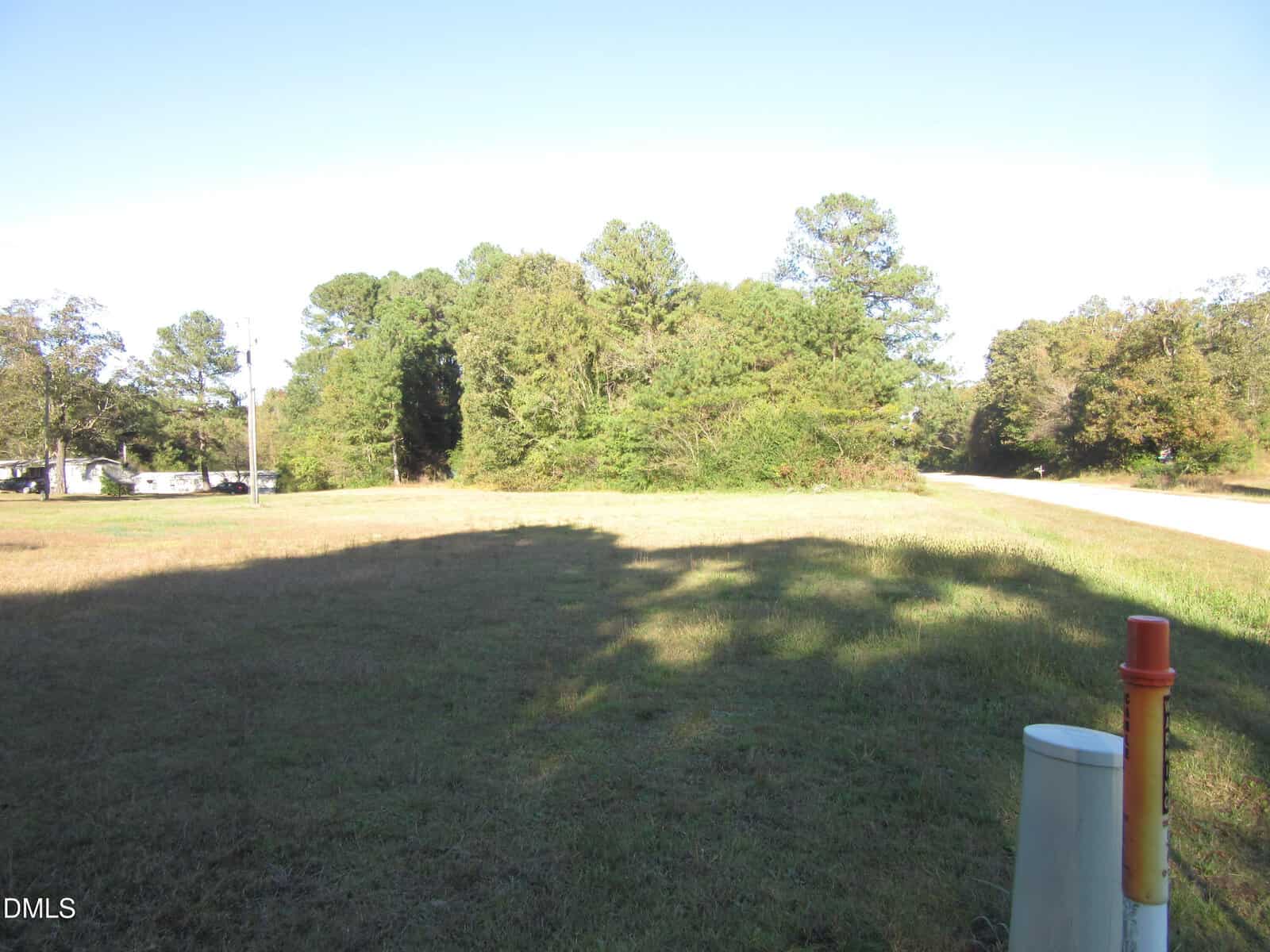 A grassy open lot with tall trees in the background and a small paved road curving to the right. Utility markers are visible in the front right corner. The sky is clear and blue.
