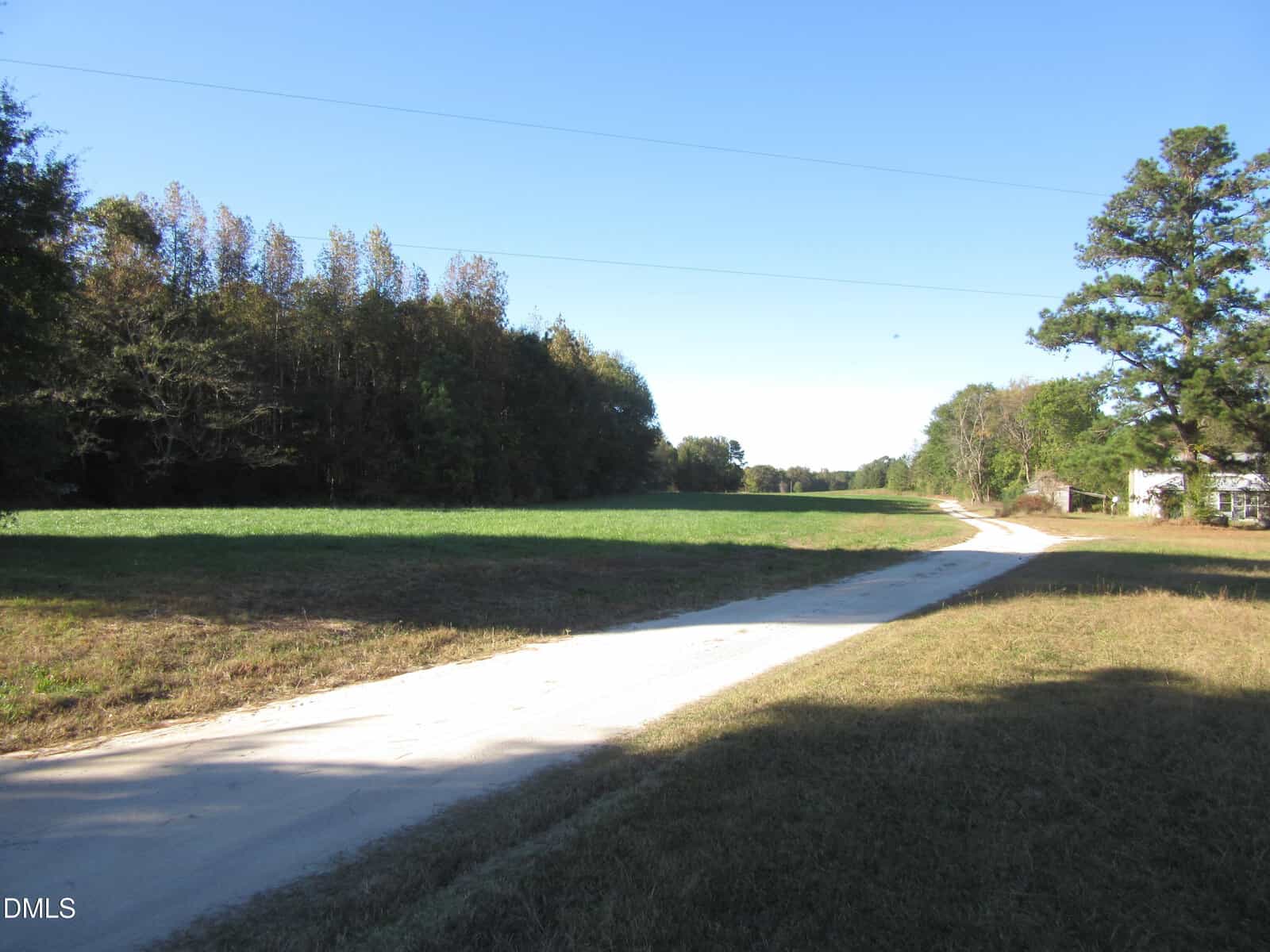 A narrow dirt road curves through a grassy field bordered by trees under a clear blue sky, with a few small buildings visible to the right.