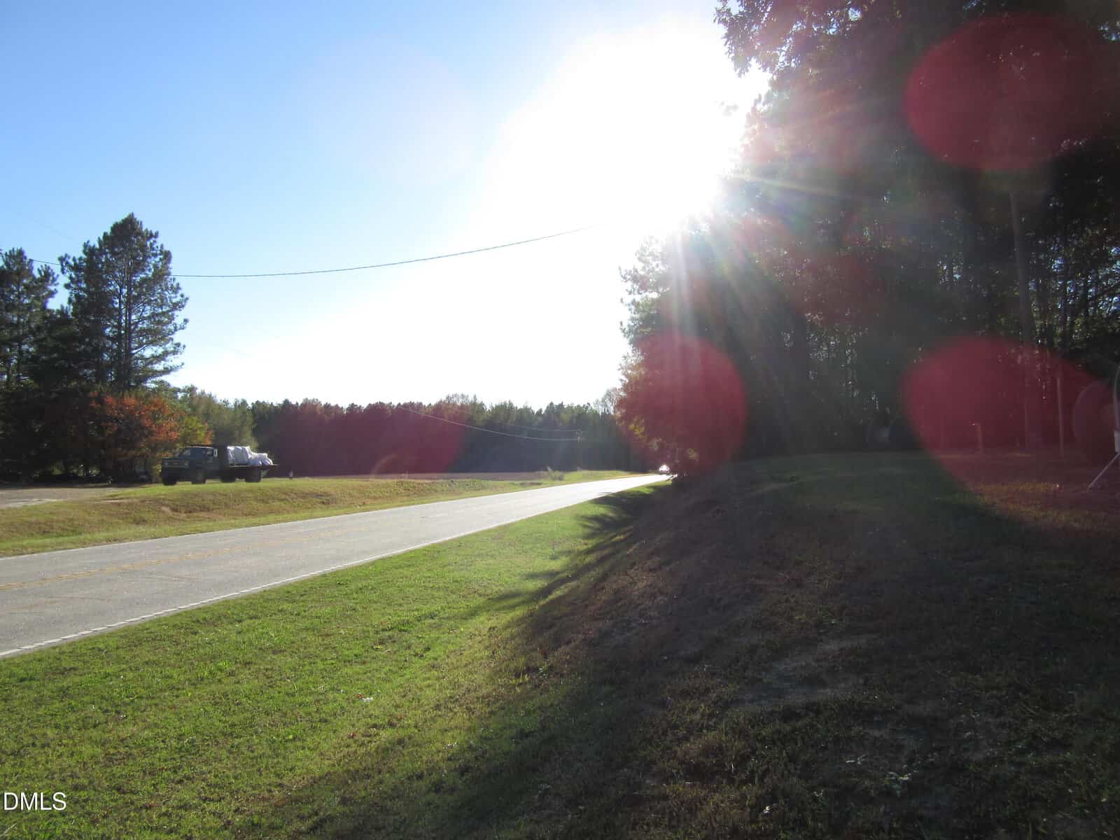 A rural road curves through grassy land bordered by trees, with sunlight streaming through the trees and creating lens flares in the image. A truck is parked near the road on the left.