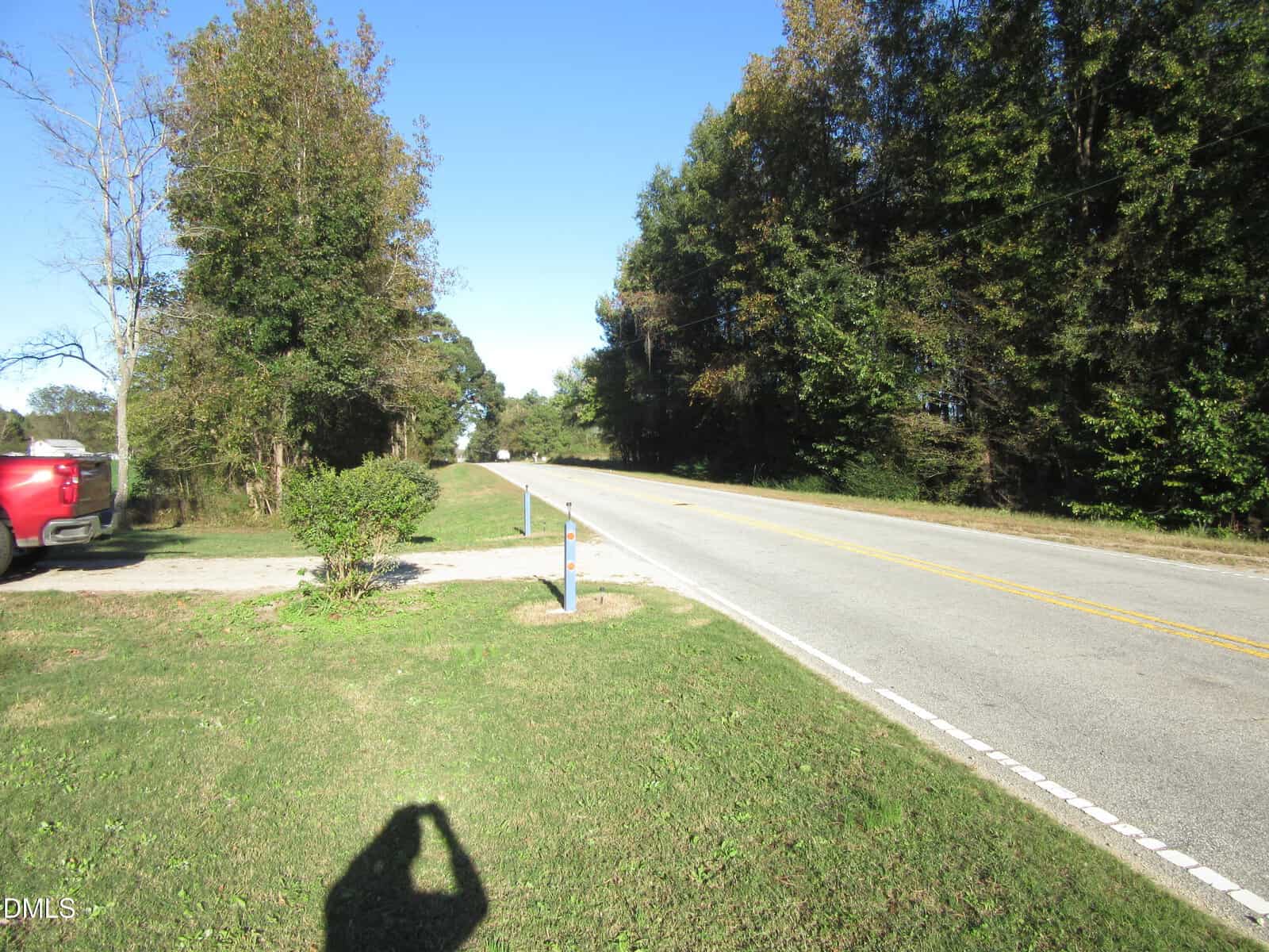 A two-lane rural road curves through a green, grassy area bordered by tall trees. A red pickup truck is parked on the left, and a person's shadow taking a photo is visible in the foreground.