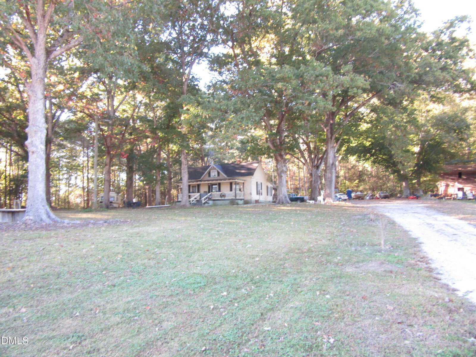A small house with a front porch sits among tall trees on a spacious grassy lot, with a dirt driveway curving toward the house and scattered items near the tree line.