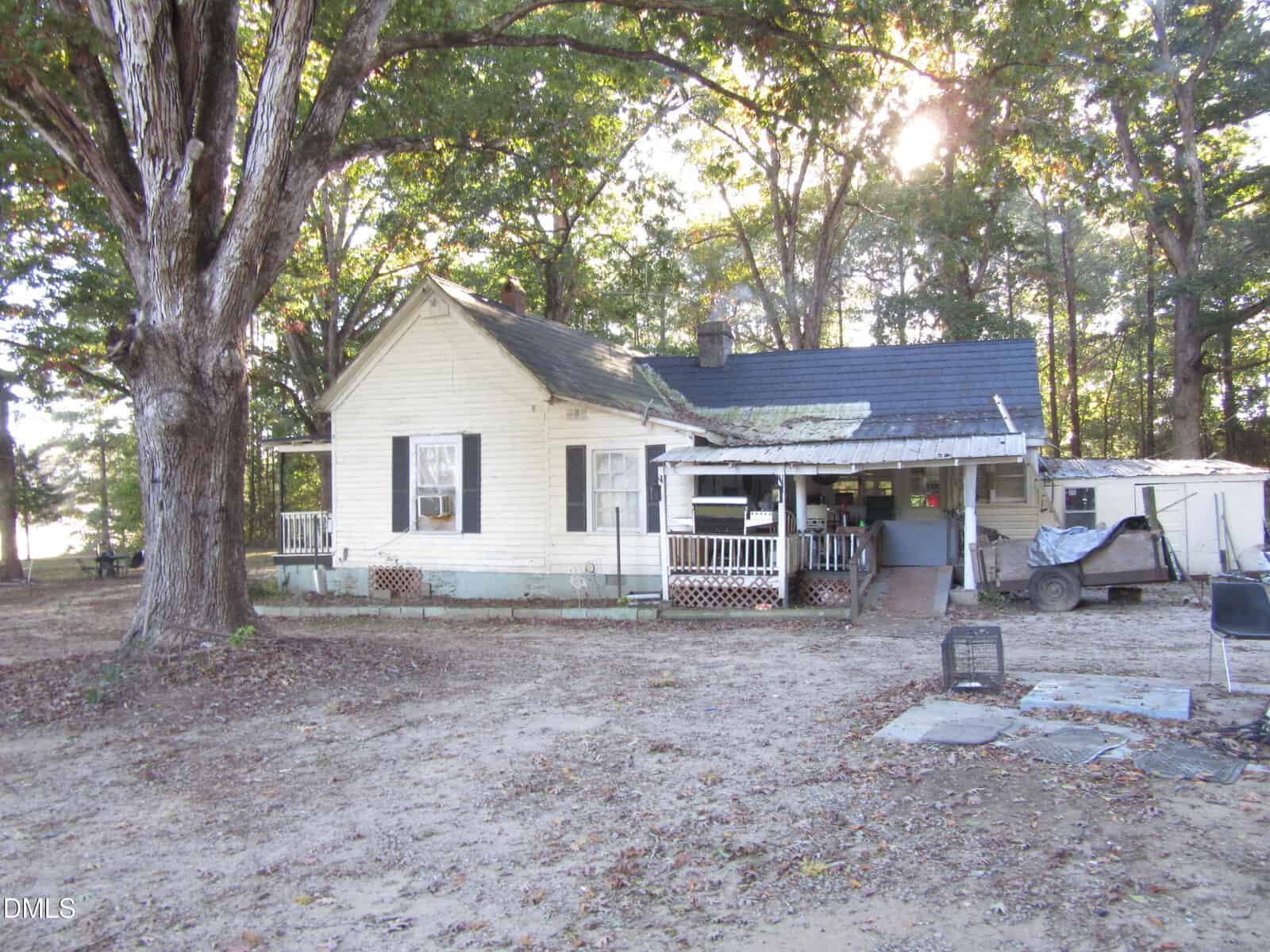 A weathered white house with a front porch sits under large trees. The yard is bare with patches of dirt and scattered items, including crates and a trailer, visible near the house. Sunlight filters through the trees.