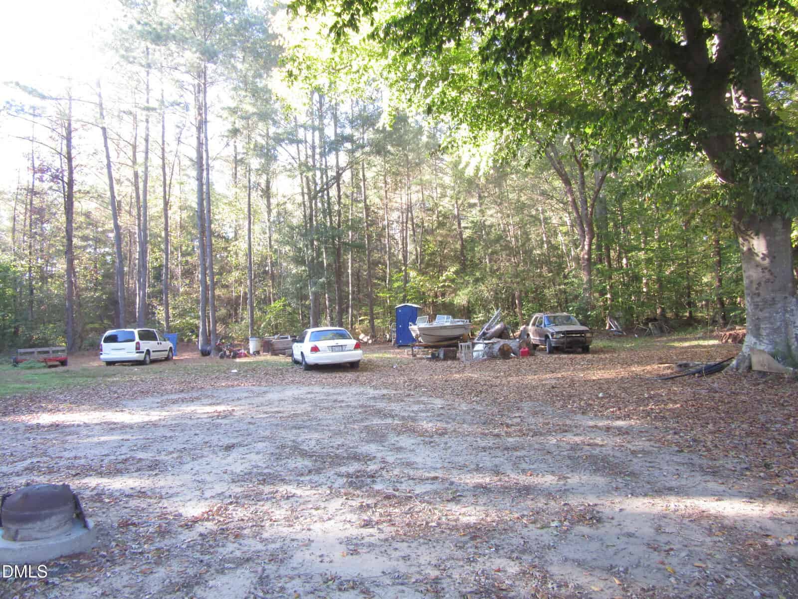 A wooded clearing with three parked cars, a boat on a trailer, and scattered items. Tall trees surround the area, and sunlight filters through the leaves onto the ground covered with dry leaves.