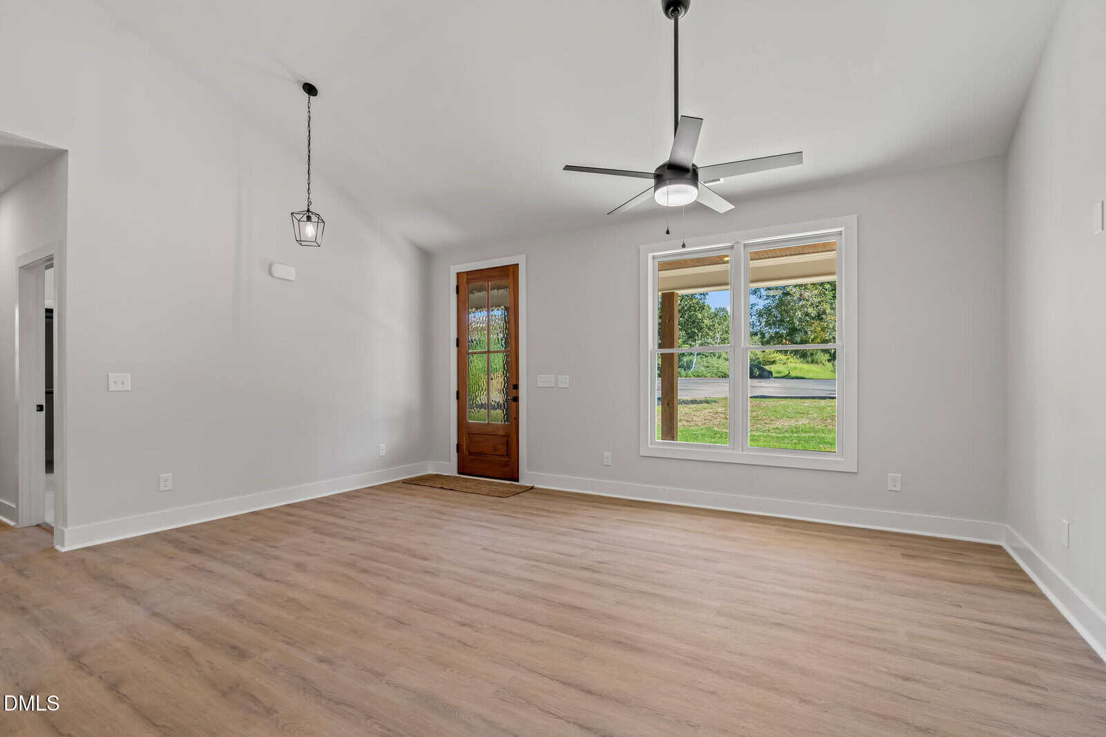 Bright, empty living room with light wood flooring, white walls, a wooden front door with glass panels, large window, ceiling fan, and hanging light fixture; view of green grass and trees outside.