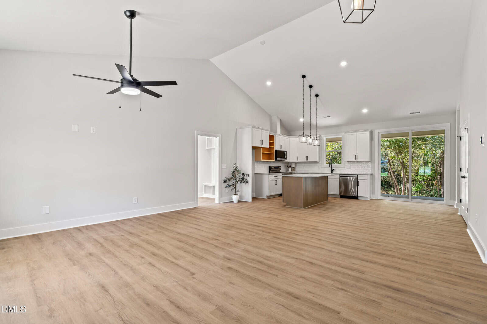 Open-concept living area with wood floors, white walls, ceiling fan, and modern kitchen featuring white cabinets, stainless steel appliances, pendant lights, and large windows overlooking greenery.