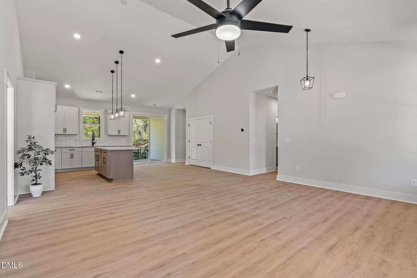 Spacious modern living area with light wood floors, white walls, a ceiling fan, and hanging lights. Open kitchen with island, white cabinets, and large windows offering outdoor views. A potted plant decorates one corner.