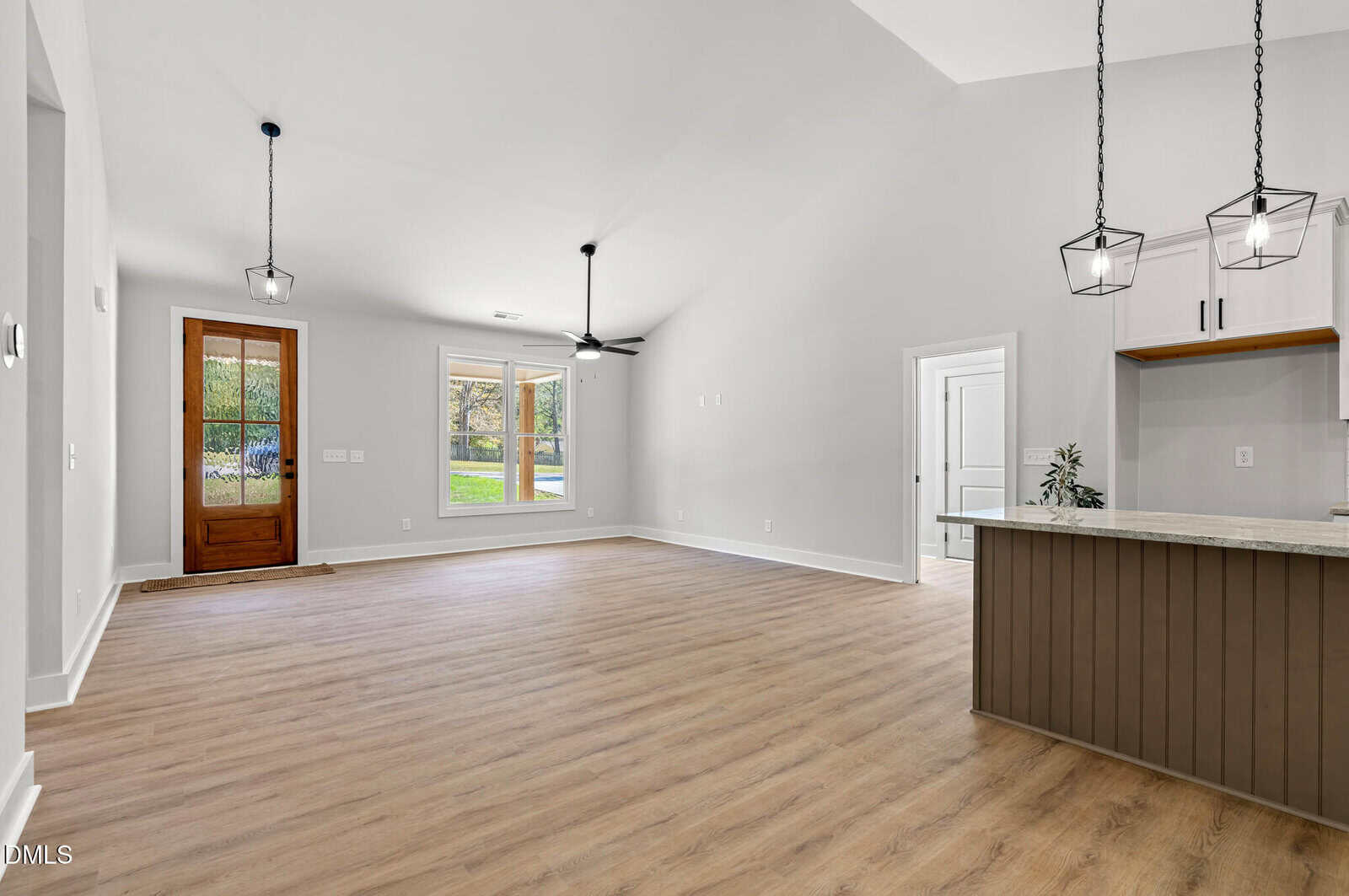 Spacious, modern open-concept living area with light wood flooring, white walls, large window, wooden front door, ceiling fan, and a kitchen with pendant lights and an island.